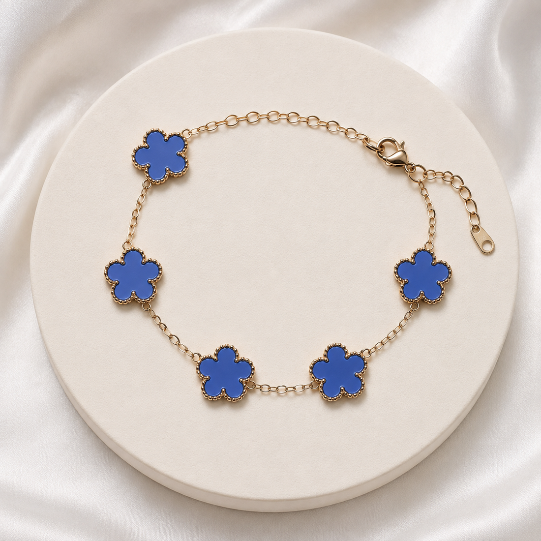 A gold chain bracelet with five blue flower-shaped links, displayed on a round cream-colored pedestal against a white satin background.